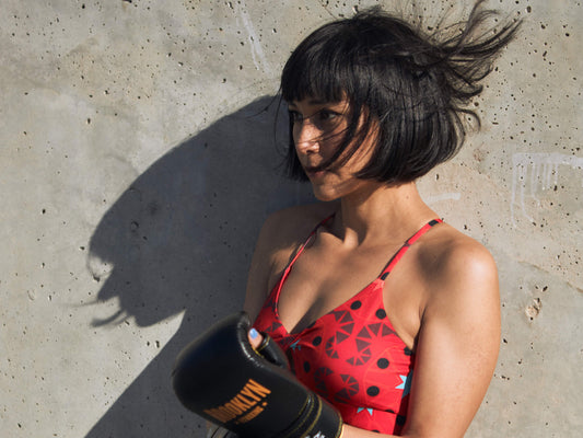 woman wearing a colorful, geometric sports bra top under a white tank top. She has her right hand over her shirt to show a bit of the sporty bra. She wears a gold bangle and is outside, in front of a cactus.