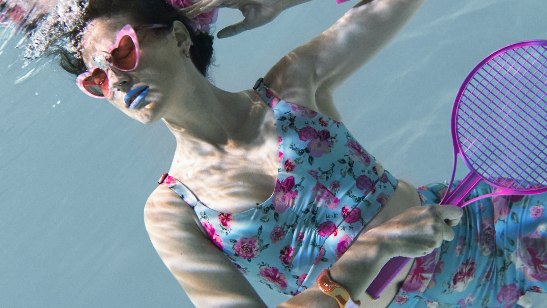 Woman underwater in a pool wearing a floral sports bra swimsuit and holding a purple badminton racket.